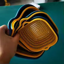 A hand holding a stack of square plastic colanders or strainers with yellow and blue rims, shown against a teal background.